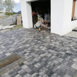Ułożona szara kostka brukowa na podjeździe przed domem z otwartym garażem; w tle widoczny pracownik w trakcie pracy, a także betonowy płotek i trawnik.