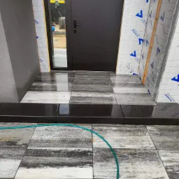 Wejście do domu z czarnymi schodami kamiennymi, ułożona szara kostka brukowa imitująca drewno, widoczny wąż ogrodowy i prace wykończeniowe elewacji.