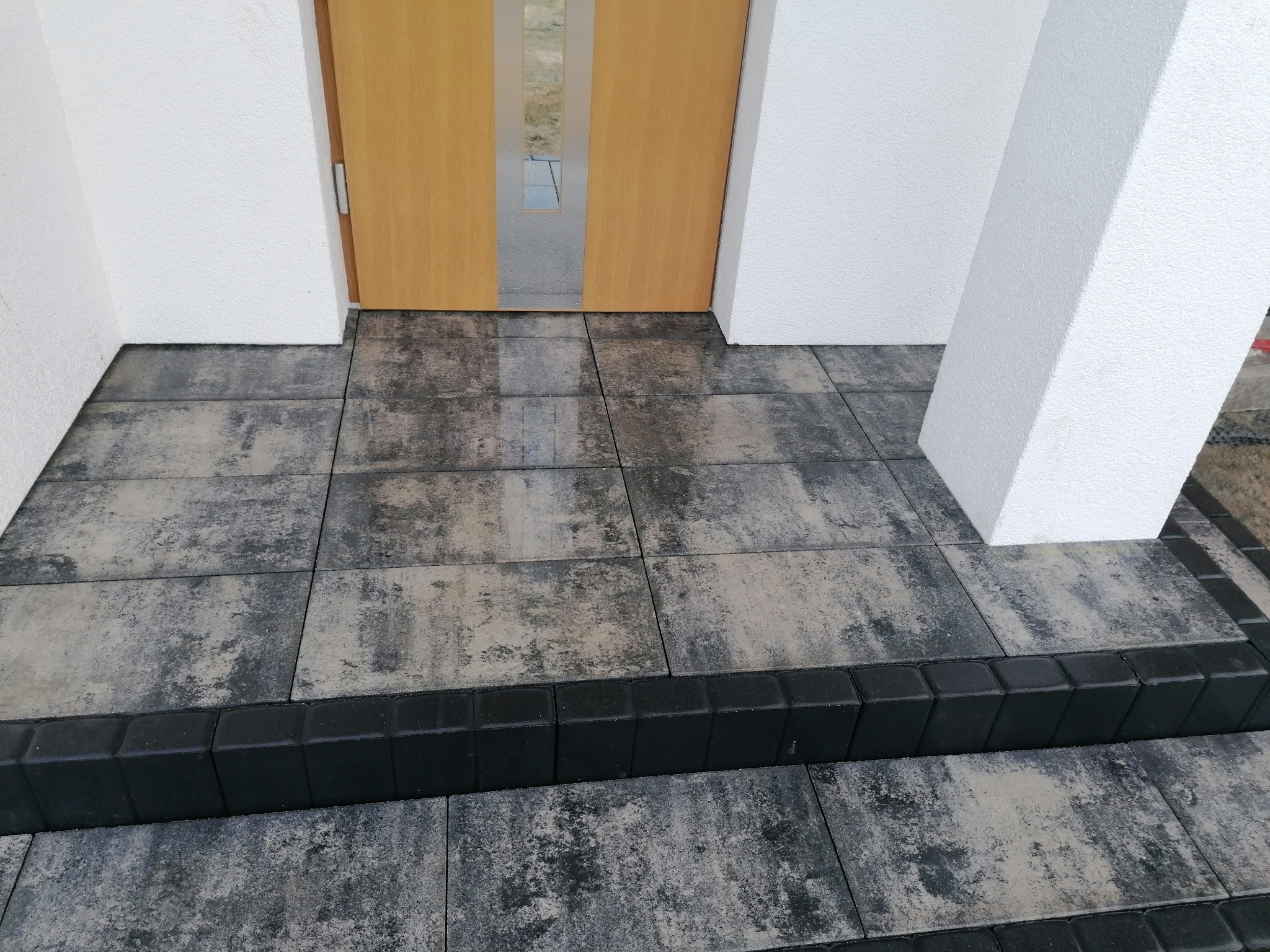 Ułożona kostka brukowa w odcieniach szarości tworząca schody wejściowe do domu z drewnianymi drzwiami, obrzeża z czarnej kostki.