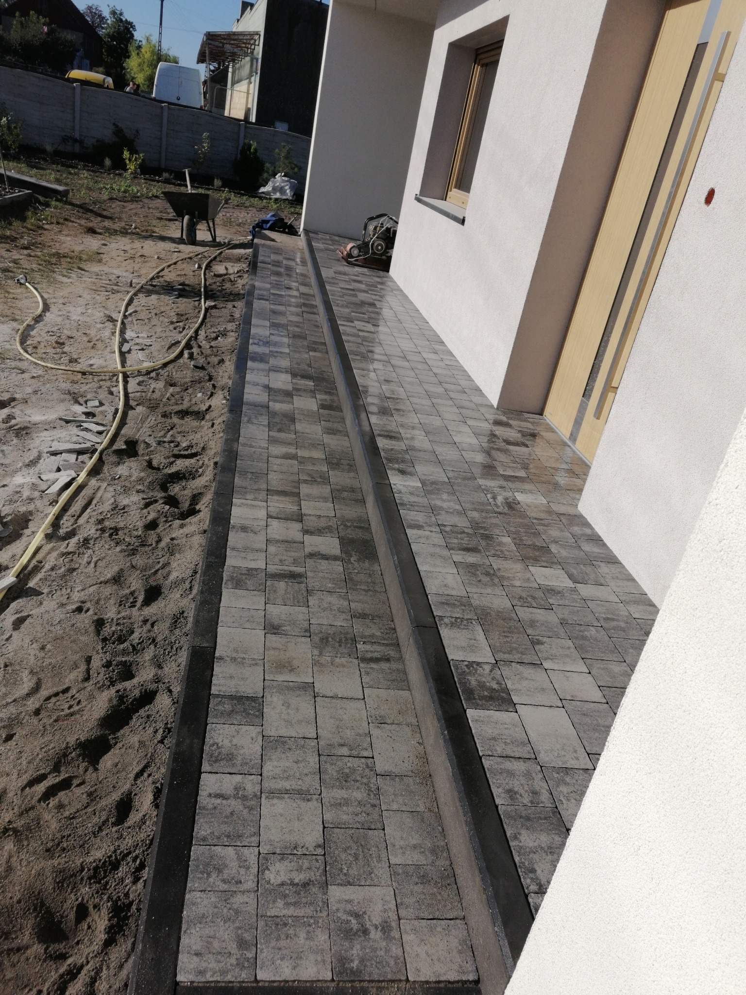 Ułożona kostka brukowa w odcieniach szarości z czarnym obramowaniem, prowadząca do drzwi wejściowych domu z widocznym wężem ogrodowym i taczką w tle.