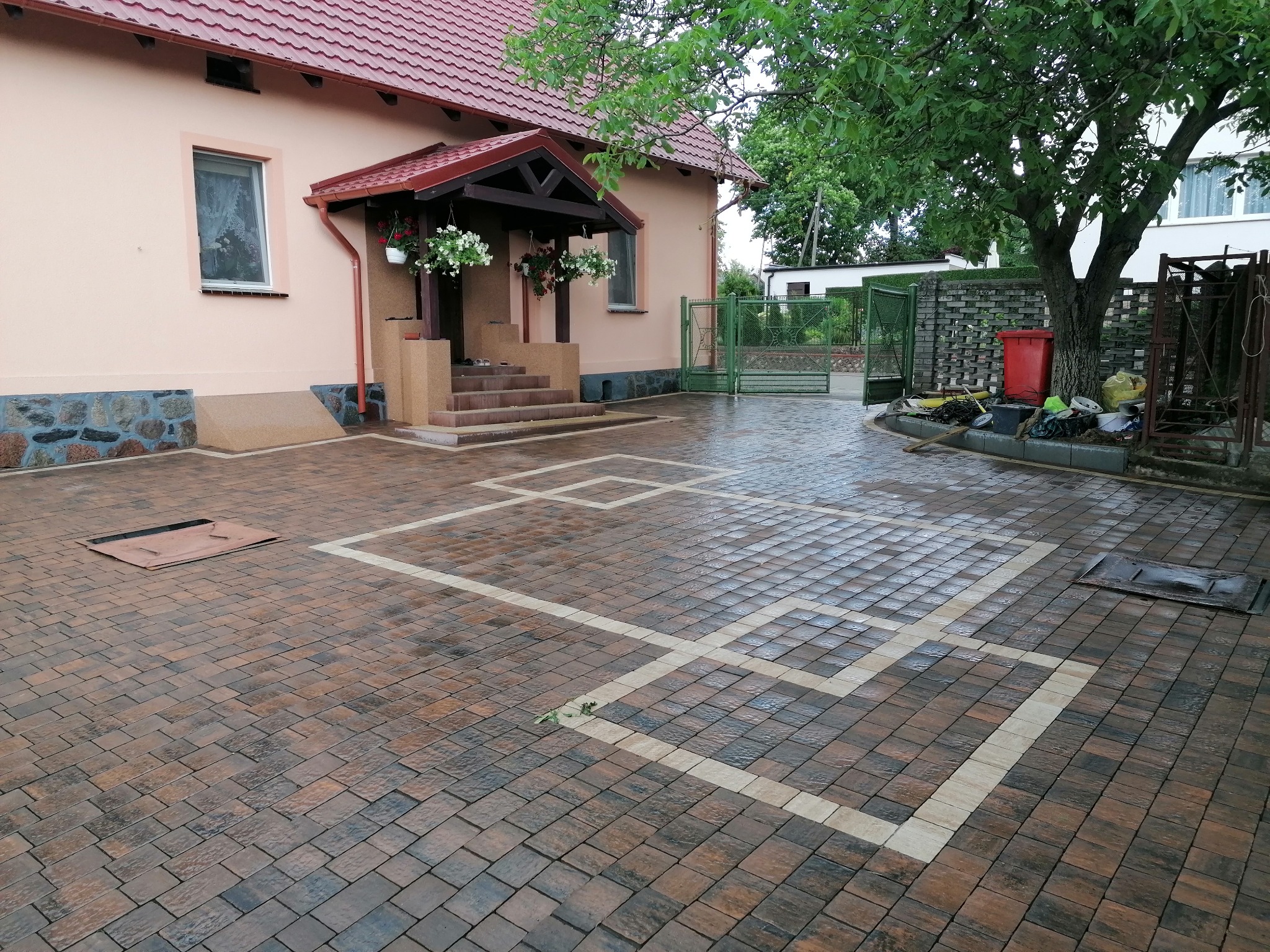 Ułożona kostka brukowa w kolorach brązu i grafitu z jasnymi liniami tworzącymi wzór przed domem z czerwonym dachem i schodami wejściowymi.