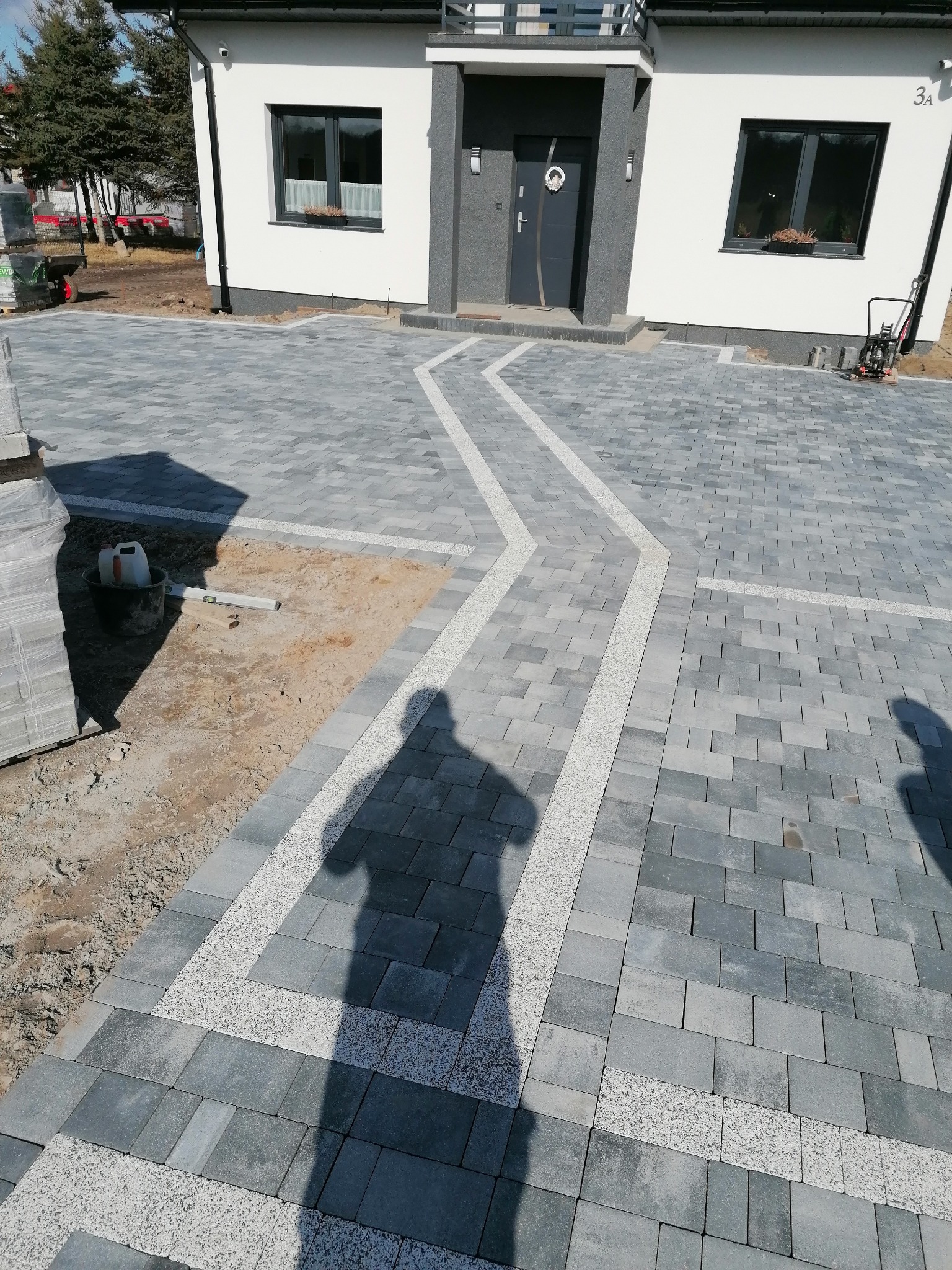 Nowoczesny podjazd z szarej kostki brukowej ułożonej we wzór, z jasnymi pasami z drobnego kamienia, prowadzący do domu z białymi ścianami i szarymi elementami architektonicznymi, widoczny cień...