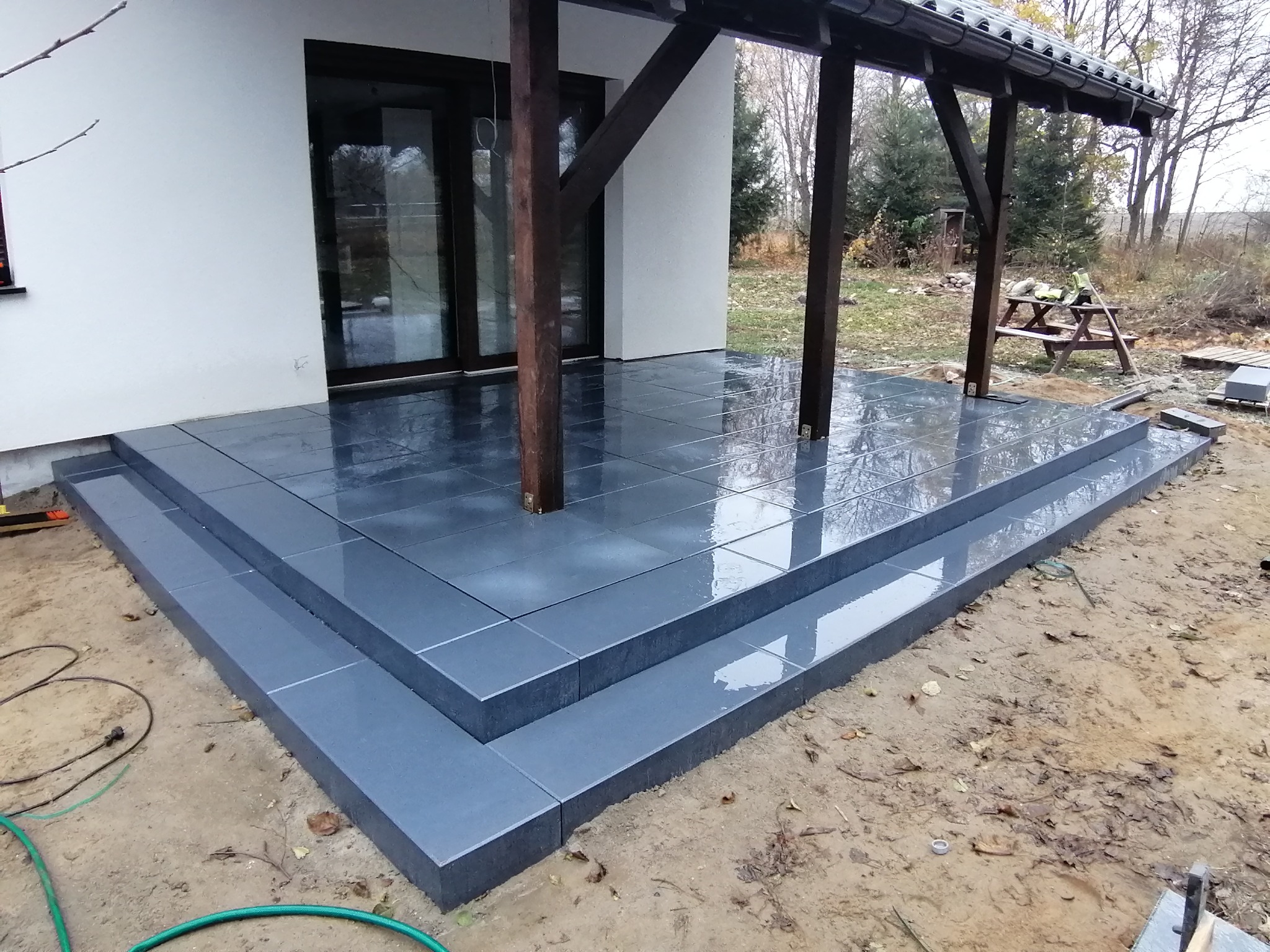 Nowoczesny taras kamienny z ciemnoszarych płyt z wyraźnymi fugami i schodami, widoczne drewniane słupy zadaszenia, teren jeszcze niezagospodarowany.