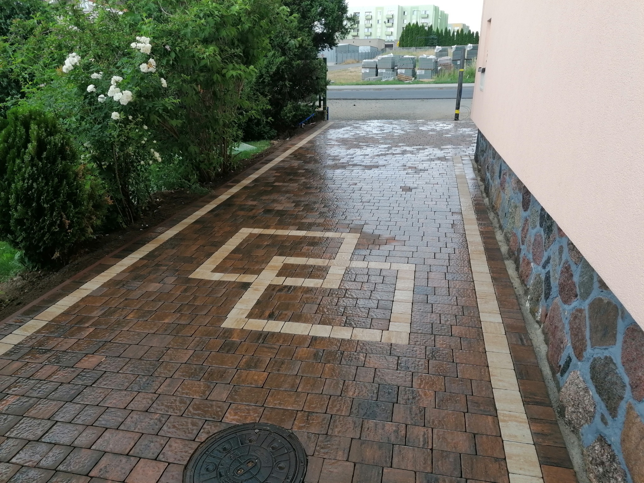 Ścieżka z brązowej kostki brukowej z jasnym geometrycznym wzorem w kształcie labiryntu, prowadząca wzdłuż ściany budynku z kamienną podmurówką i rabatą z kwitnącymi krzewami.