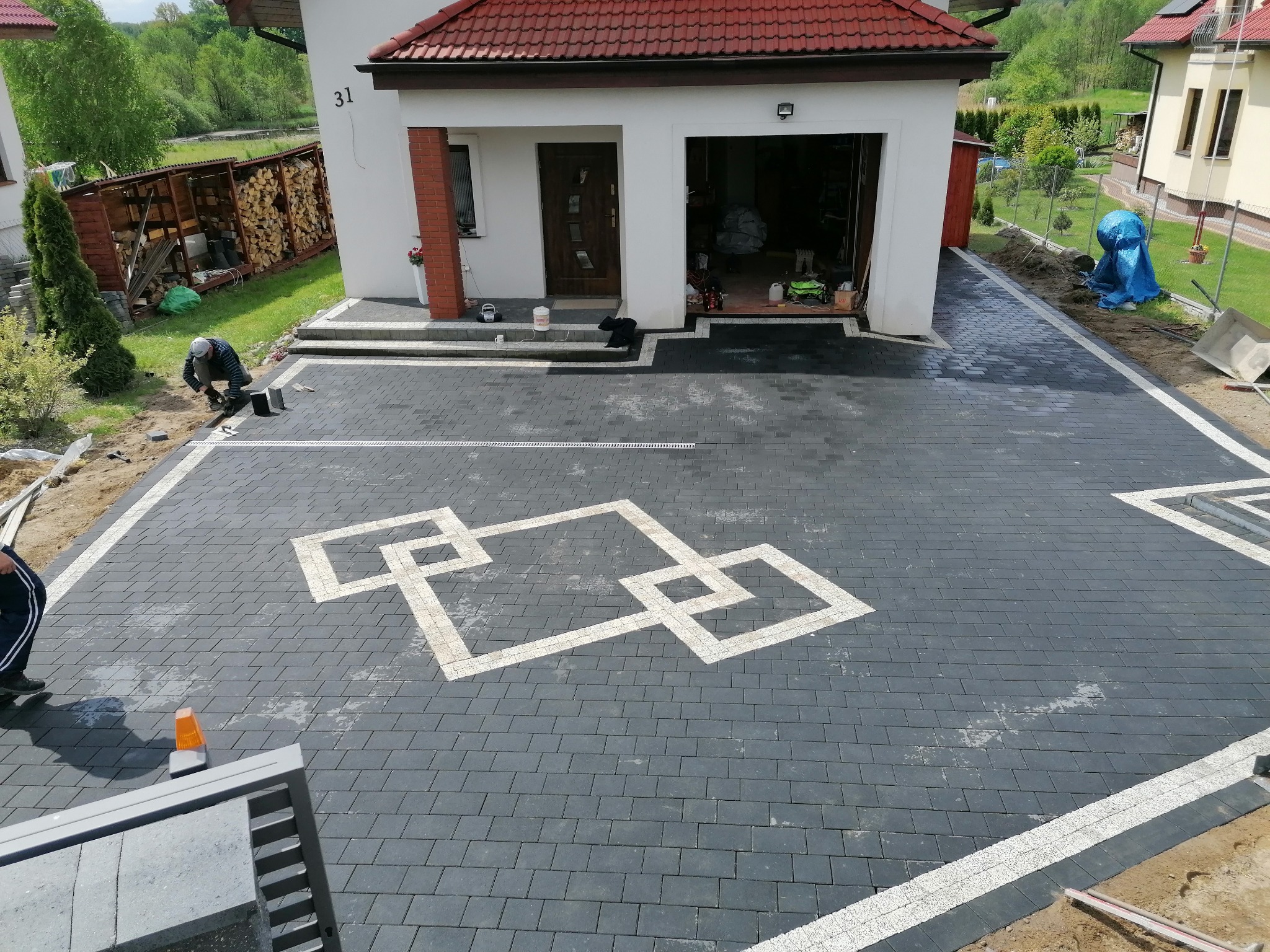 Nowo ułożony podjazd z ciemnoszarej kostki z dekoracyjnym wzorem z jasnej kostki, widoczny pracownik podczas układania kostki oraz dom z garażem w tle.