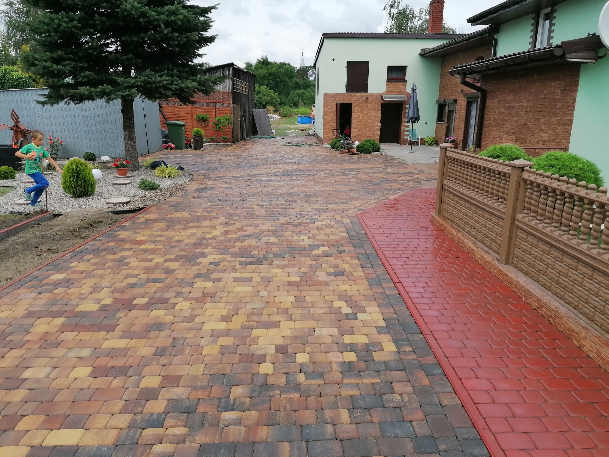 Szeroki, wzorzysty podjazd z kostki brukowej w odcieniach brązu, żółci i szarości, z czerwonym obramowaniem z kostki, prowadzący do domu z ogrodem, gdzie biega dziecko.