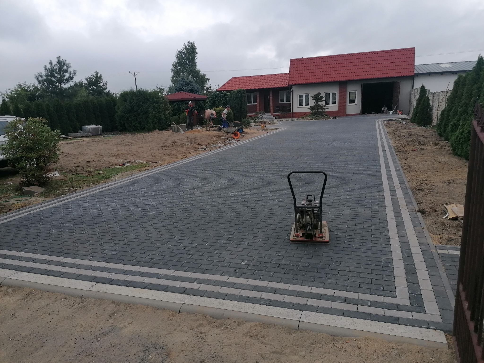 Ułożona, szara kostka brukowa z jasnymi pasami po bokach, zagęszczarka wibracyjna na powierzchni, w tle dom z czerwoną dachówką i pracujący robotnicy.