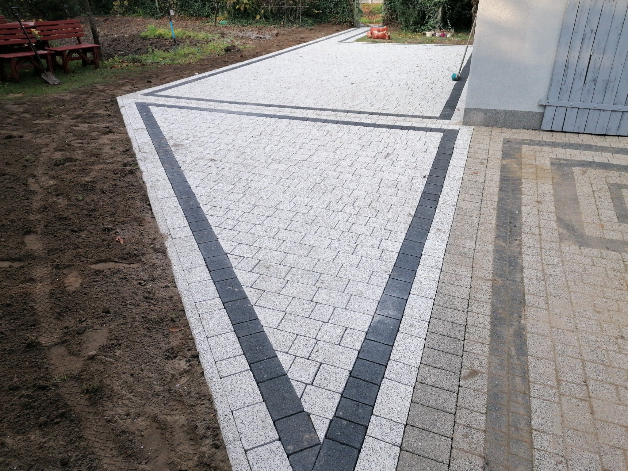 Wykończona nawierzchnia z jasnoszarej kostki brukowej z ciemnoszarą obwódką, prowadząca od trawnika i drewnianej ławki do budynku z szarą bramą.