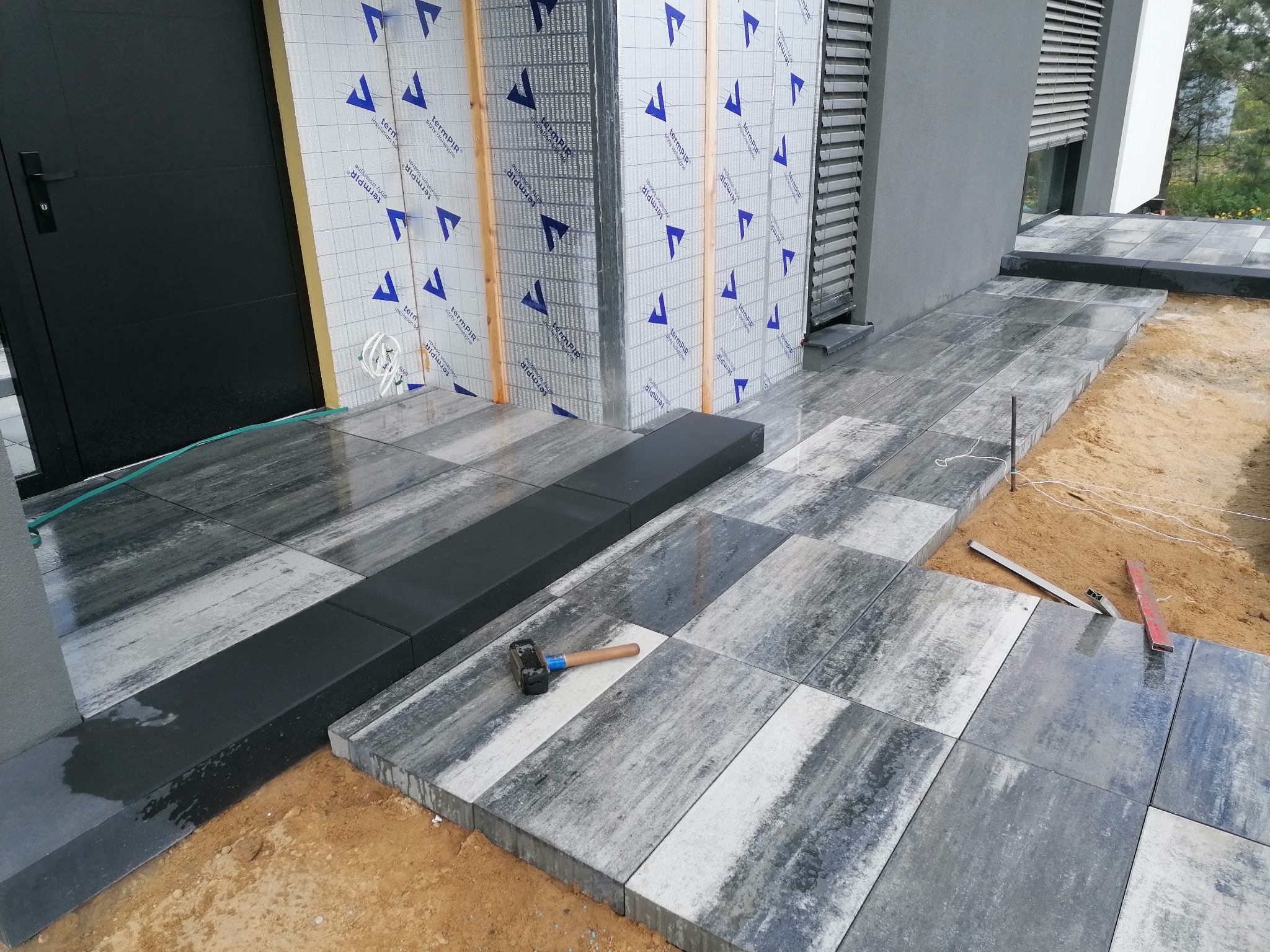 Fragment układanej ścieżki z kostki brukowej w Trzemesznie, widoczny gumowy młotek i poziomica, tło stanowi elewacja budynku z ociepleniem.