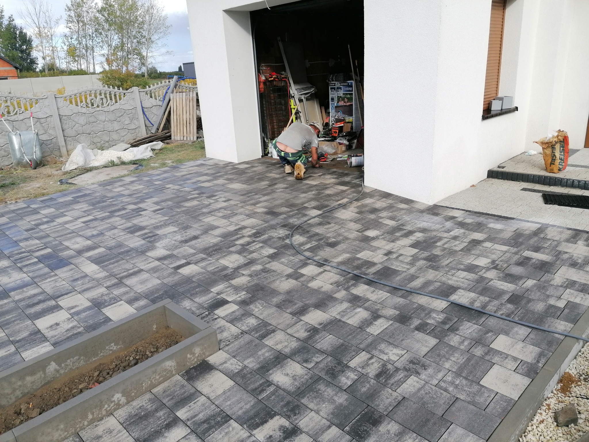 Ułożona szara kostka brukowa na podjeździe przed domem z otwartym garażem; w tle widoczny pracownik w trakcie pracy, a także betonowy płotek i trawnik.