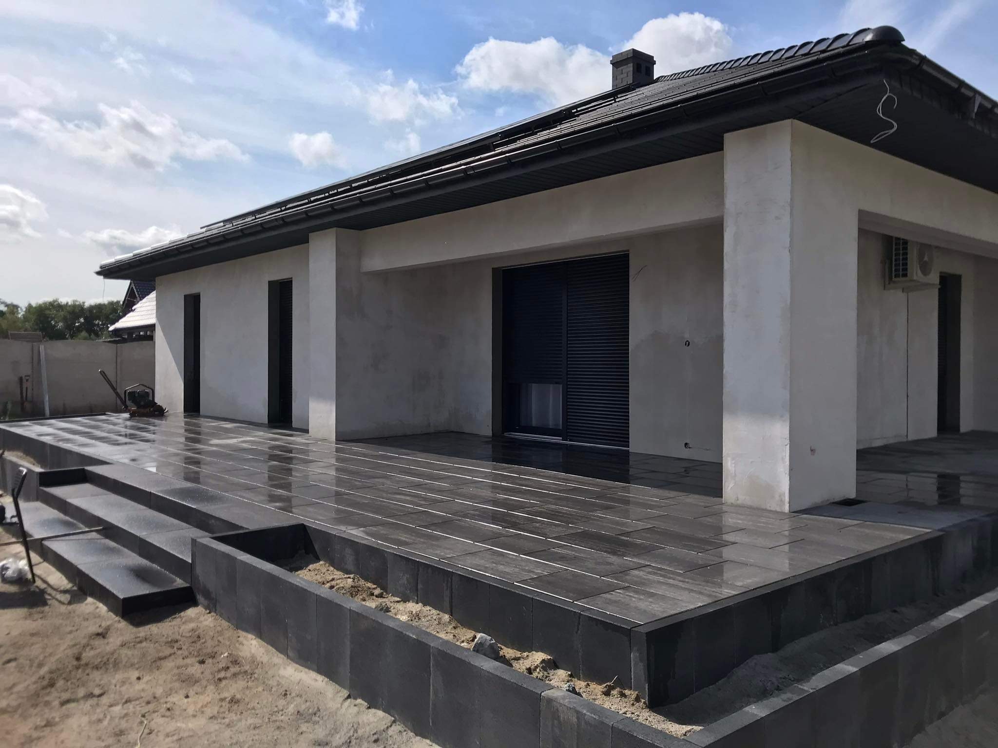 Nowoczesny taras z ciemnoszarej kostki brukowej, prowadzący do domu z jasną elewacją, widoczne trzy stopnie i obrzeże z kostki, teren wokół w trakcie prac ziemnych.