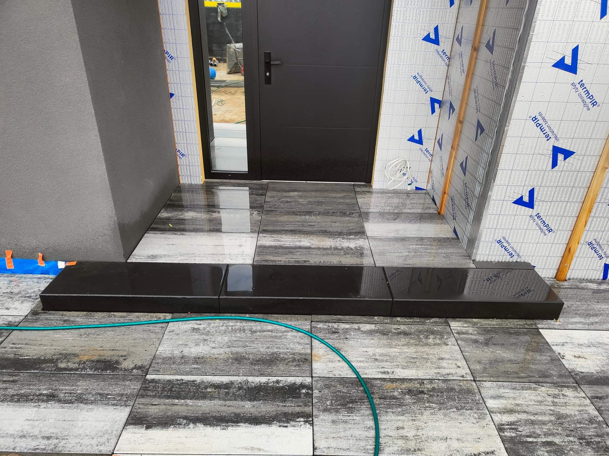 Wejście do domu z czarnymi schodami kamiennymi, ułożona szara kostka brukowa imitująca drewno, widoczny wąż ogrodowy i prace wykończeniowe elewacji.