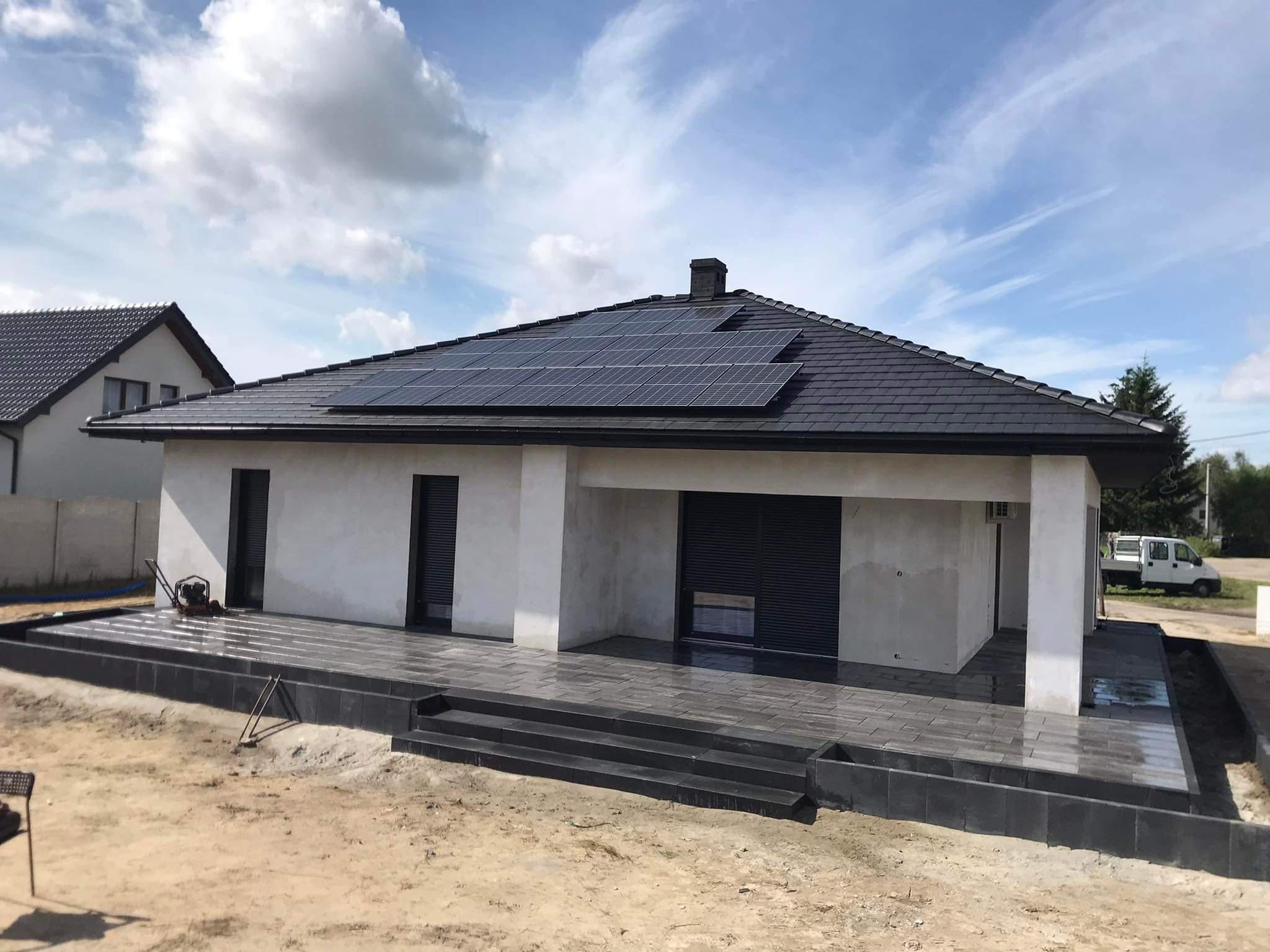 Nowoczesny dom z szarego kamienia z tarasem i schodami z ciemnej kostki, panele słoneczne na dachu, widok na plac budowy.