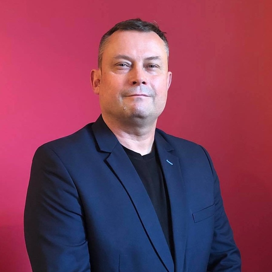 Portret mężczyzny w granatowej marynarce i czarnym T-shircie na tle czerwonej ściany z delikatnym gradientem.