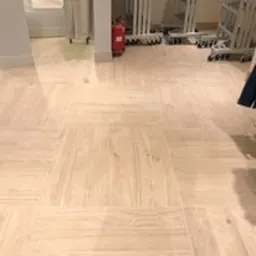 Jasna, drewnopodobna podłoga z płytek ułożonych w kwadraty, z widocznym fragmentem ściany, gaśnicą i metalowymi wózkami w tle. Płytki o delikatnej strukturze drewna, ułożone naprzemiennie, tworzą...