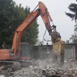 Pomarańczowa koparka Doosan z hydraulicznymi nożycami do betonu rozbiera murowany budynek. Widoczne gruz i pył.