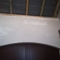 Świeżo nałożona, jasnoszara warstwa tynku na ścianie nad brązowymi drzwiami, z widoczną konstrukcją drewnianą sufitu i fragmentami ceglanego wykończenia po bokach.