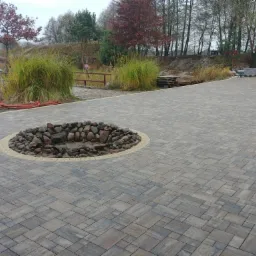 Ułożona kostka brukowa w odcieniach szarości i brązu z okrągłym elementem dekoracyjnym z kamieni na środku, w tle widoczna zieleń i drzewa.