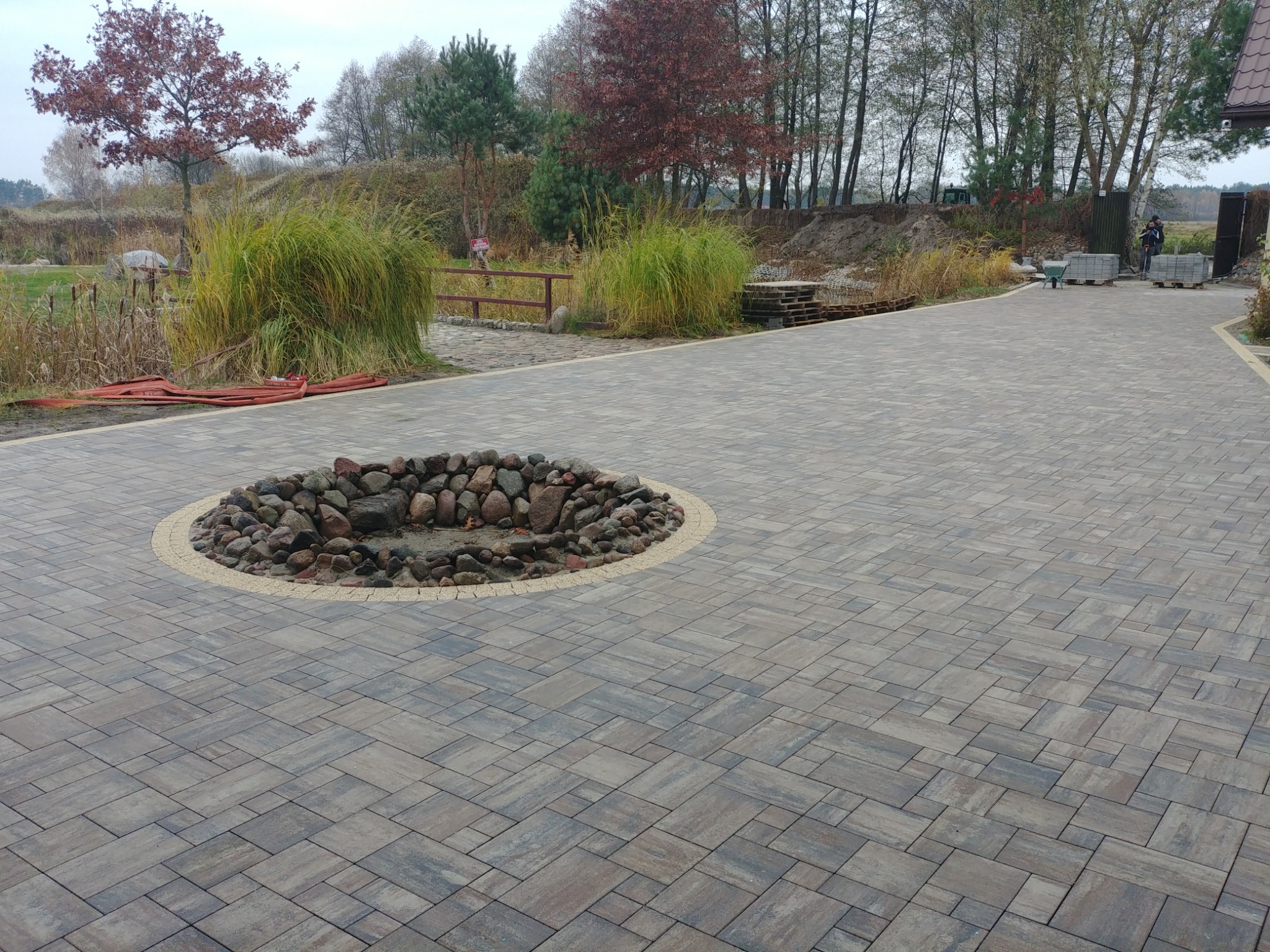 Ułożona kostka brukowa w odcieniach szarości i brązu z okrągłym elementem dekoracyjnym z kamieni na środku, w tle widoczna zieleń i drzewa.