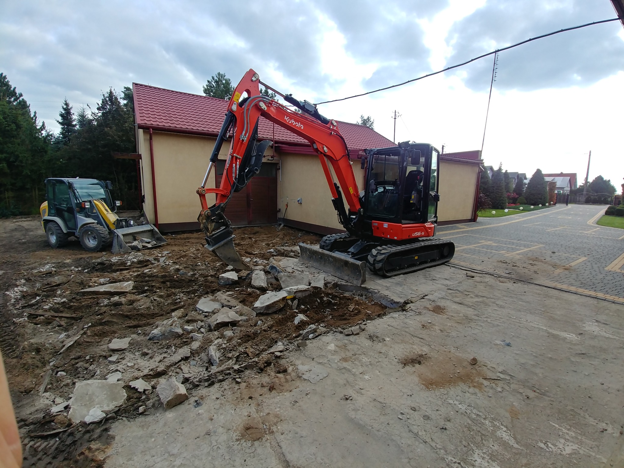 Czerwona mini koparka Kubota w trakcie prac ziemnych na posesji, z gruzem i wykopanym podłożem, w tle budynek z czerwoną dachówką i mała ładowarka.