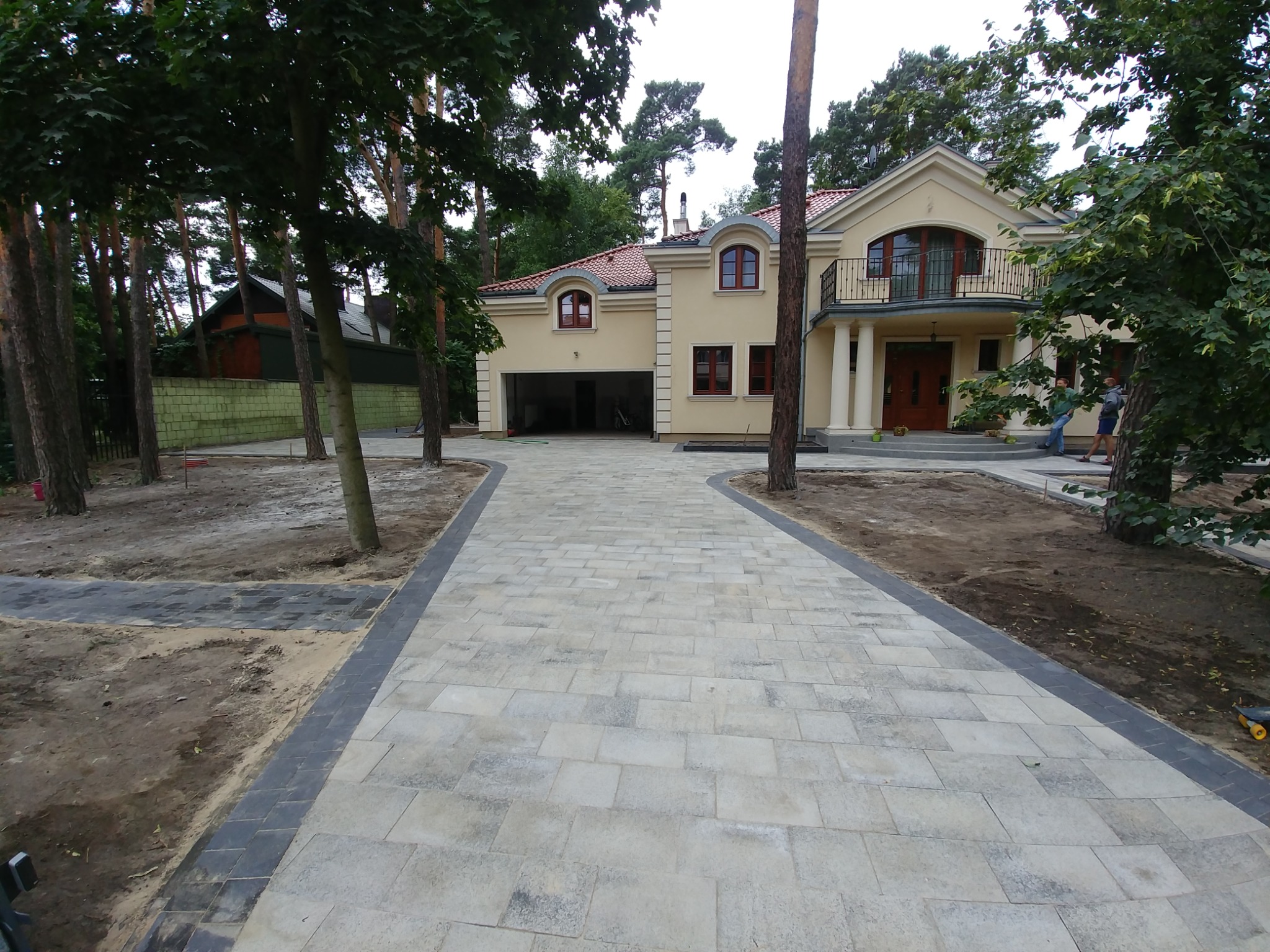 Szeroki podjazd z jasnej kostki brukowej prowadzący do beżowego domu z garażem, obrzeża z ciemnej kostki, drzewa po bokach, częściowo wykończony teren.