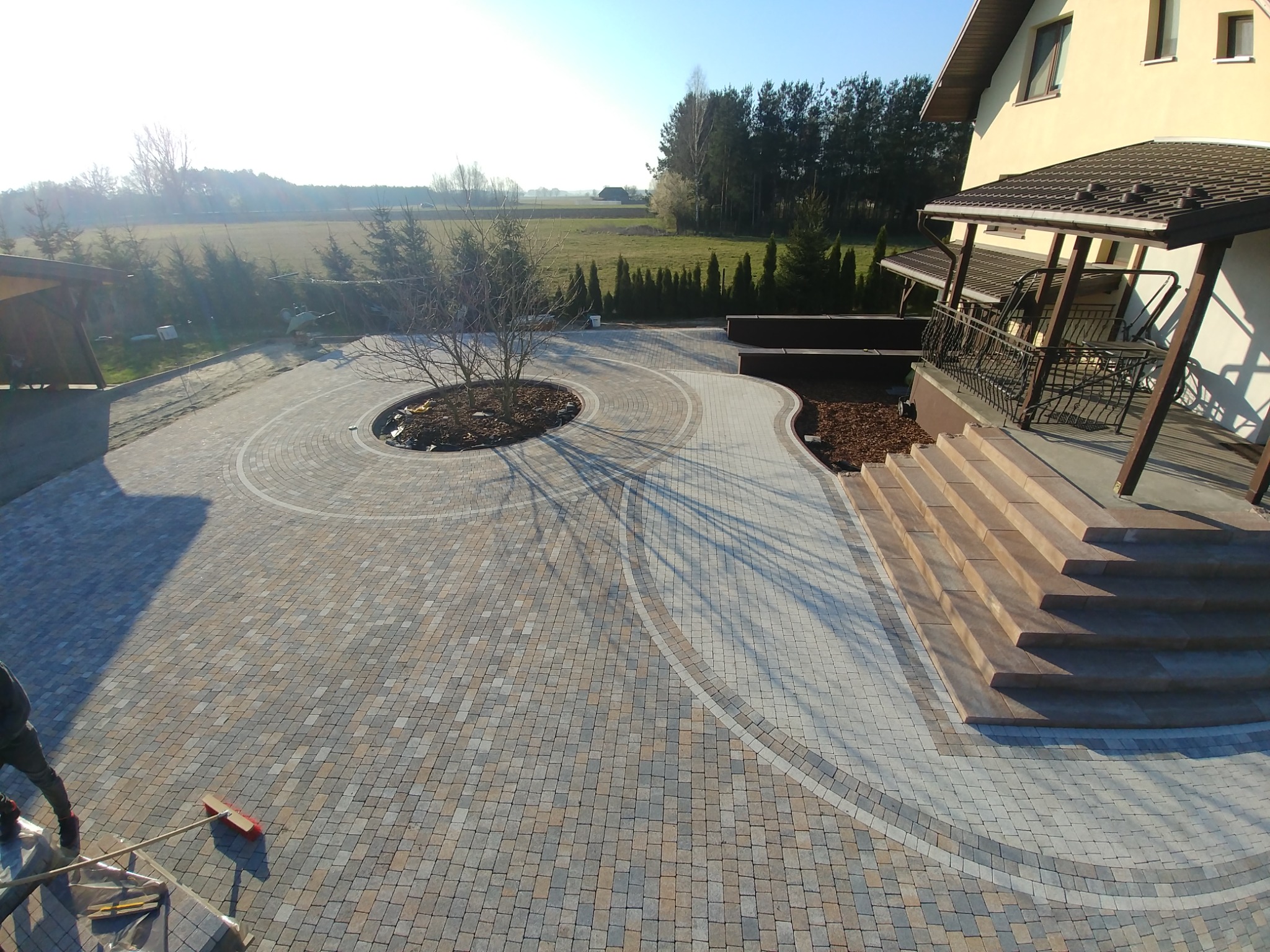 Szeroki, wybrukowany podjazd z okrągłym klombem z drzewem pośrodku, prowadzący do schodów wejściowych domu z balkonem, widok z góry, częściowo w cieniu, z widocznym fragmentem pracownika z miotłą...