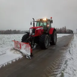 Czerwony traktor Massey Ferguson z pługiem odśnieżnym usuwa śnieg z asfaltowej drogi w pochmurny dzień. Widoczne ślady opon na mokrej nawierzchni i resztki śniegu na poboczu.