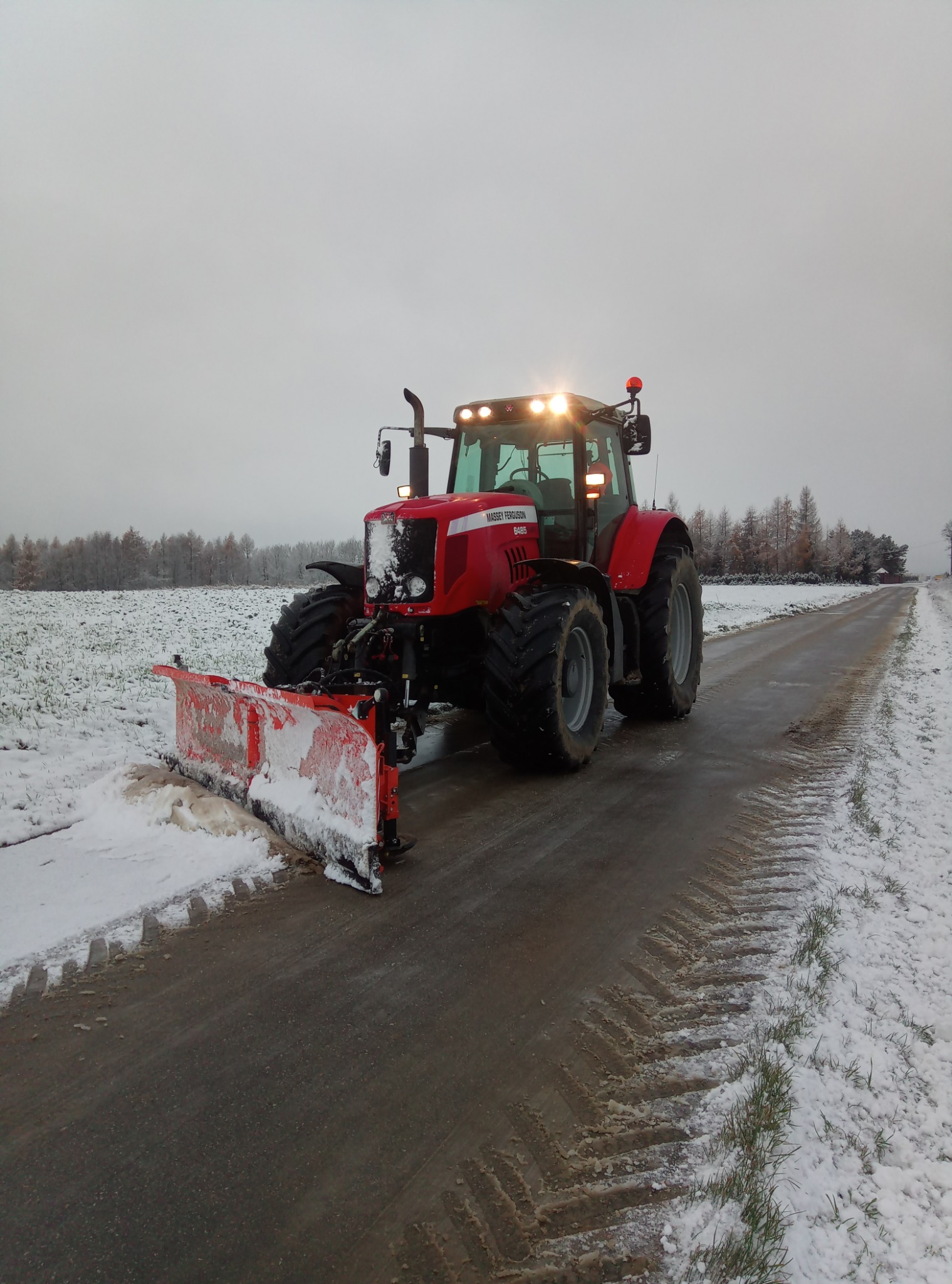 Czerwony traktor Massey Ferguson z pługiem odśnieżnym usuwa śnieg z asfaltowej drogi w pochmurny dzień. Widoczne ślady opon na mokrej nawierzchni i resztki śniegu na poboczu.