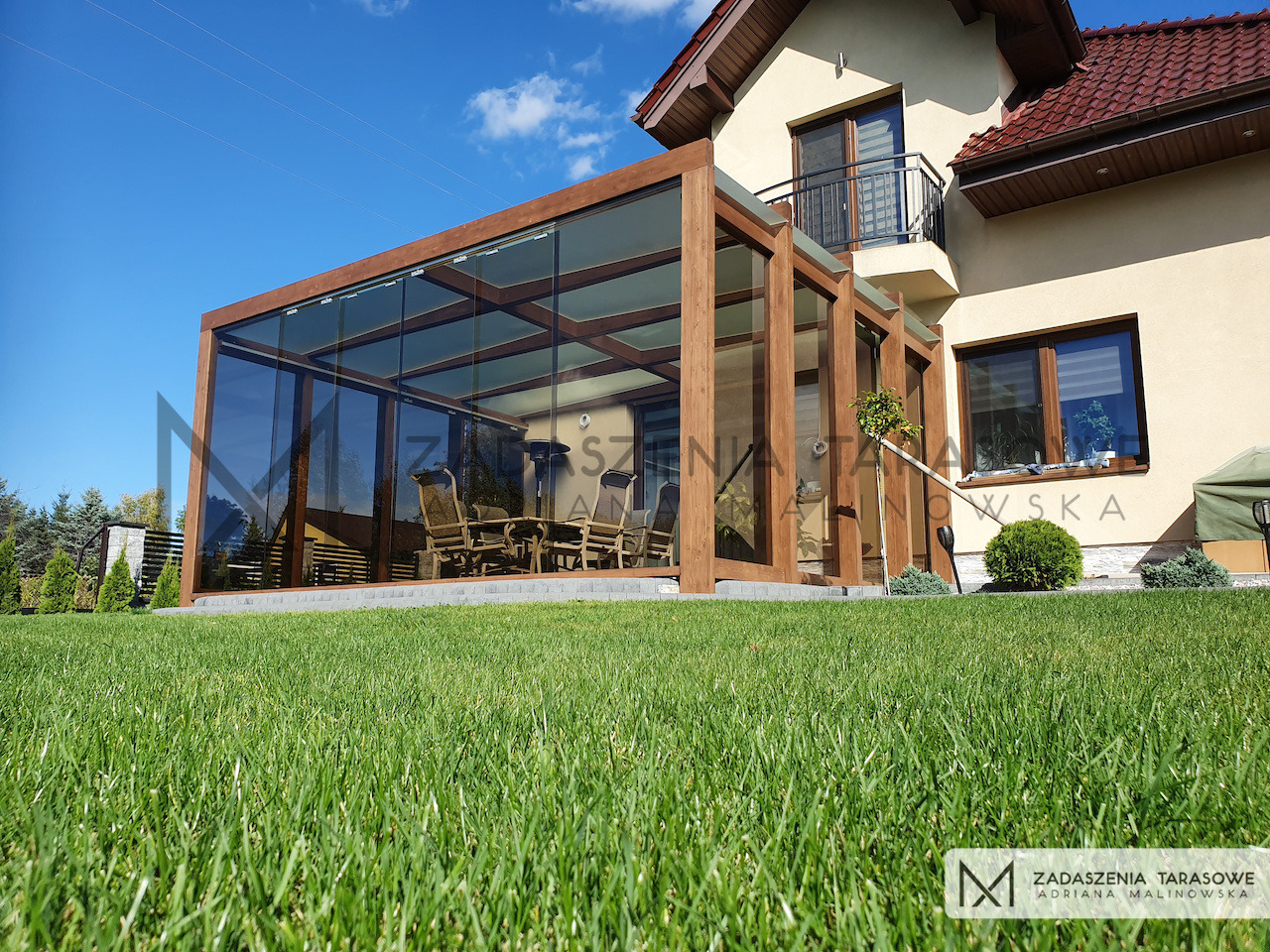 Zadaszenie tarasu ze szkłem bezpiecznym, orzechowe drewniane słupy, widok z trawnika na przydomowy ogród.