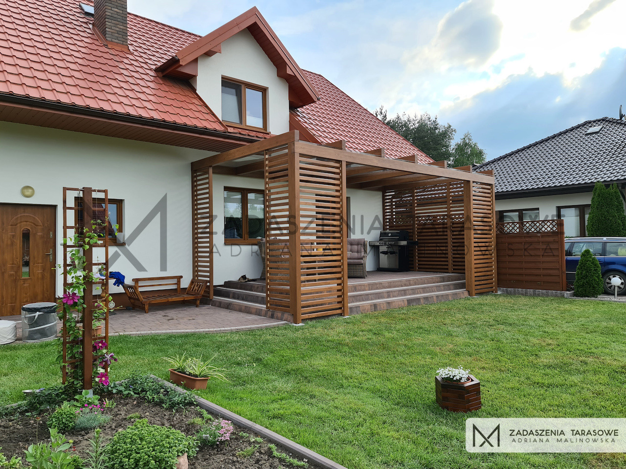 Zadaszony taras z drewnianą konstrukcją i ekranami przeciwsłonecznymi, wyposażony w grill i meble ogrodowe, przylegający do domu z czerwonym dachem.