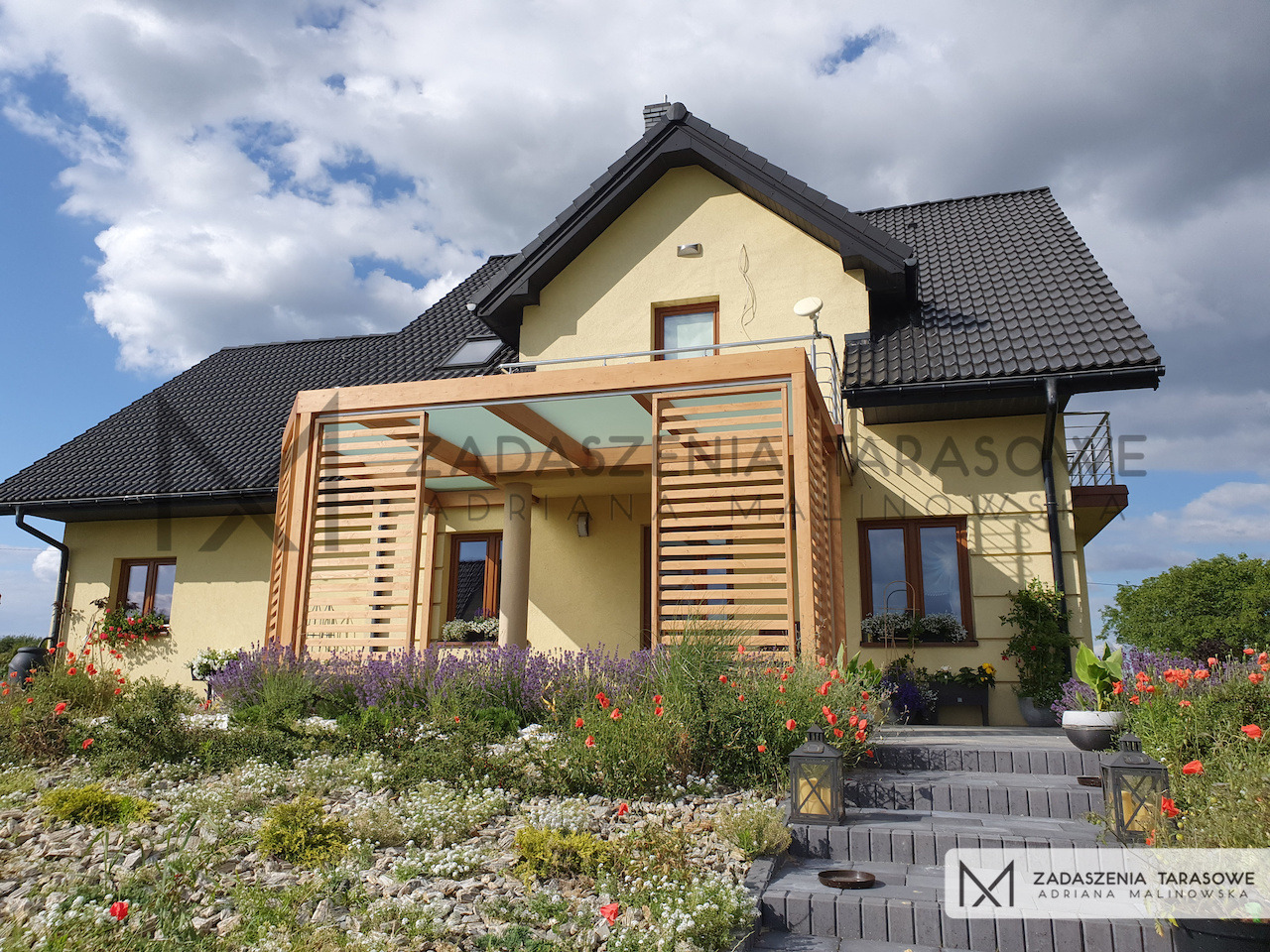 Zabudowa balkonu z drewnianą pergolą ze stałymi i przesuwnymi ekranami, wkomponowana w bryłę domu o żółtej elewacji i czarnym dachu, widok z ogrodu z kamienną ścieżką i kwitnącymi kwiatami.