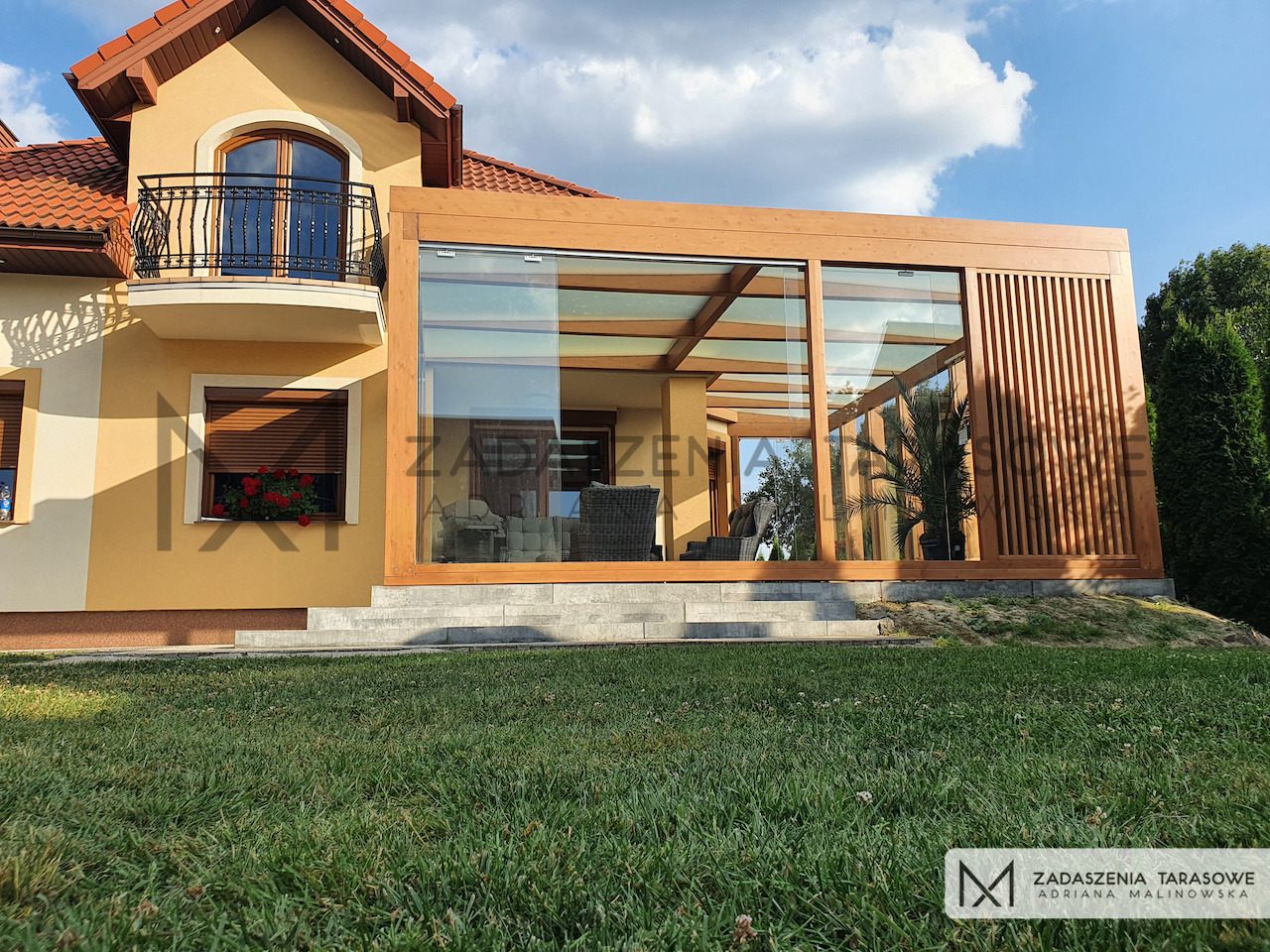 Zadaszony taras z drewnianą konstrukcją, szklanym dachem i pionowymi lamelami, przylegający do domu z balkonem.