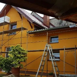 Ż&oacute;łta elewacja domu jednorodzinnego w trakcie ocieplania, widoczne rusztowania, styropian EPS na rusztowaniu i drabina oparta o ścianę, rośliny w donicach przed budynkiem.