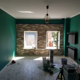 Wnętrze pokoju z zielonymi ścianami, imitacją cegły na ścianie, dwoma oknami, wiszącą lampą w stylu industrialnym i rozłożonymi narzędziami oraz materiałami budowlanymi podczas prac wykończeniowych.