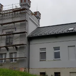 Szara elewacja budynku w stylu zamkowym z wieżyczkami, częściowo pokryta rusztowaniem, świeżo po tynkowaniu, z widocznymi śladami prac na dolnej partii ściany.