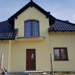 Żółty dom jednorodzinny z ciemną dachówką, brązowymi oknami i drzwiami, widoczne prace wykończeniowe przy ogrodzeniu i otwarty garaż z osobami w środku.