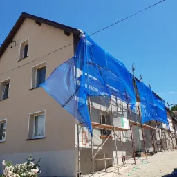 Dwupiętrowy dom w trakcie prac elewacyjnych, osłonięty niebieską siatką na rusztowaniach, widoczne fragmenty ocieplenia i świeżego tynku, na ścianie zamontowana antena satelitarna, w tle sąsiednie...