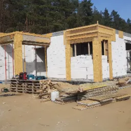 Budowa parterowego domu z białych bloczków, widoczne drewniane szalunki okienne i nadproża, palety z bloczkami Prefabet i narzędzia na budowie w słoneczny dzień.