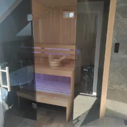 Wnętrze nowocześnie wykończonej kabiny z drewna z przeszklonymi drzwiami, widoczne drewniane ławki, piec z kamieniami Harvia, wiadro z chochlą i sterownik na zewnątrz.