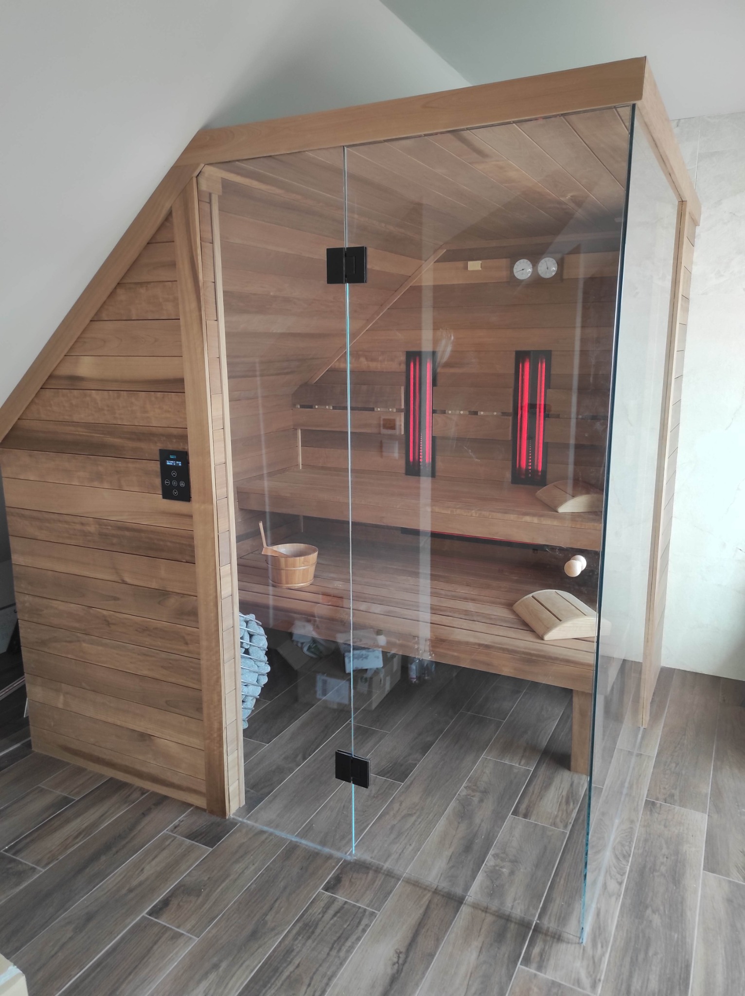 Drewniana kabina z przeszklonymi drzwiami, wnętrze z dwoma rzędami ławek, oparciami, piecem z kamieniami, oświetleniem i sterowaniem na zewnątrz, usytuowana w pomieszczeniu ze skośnym sufitem.