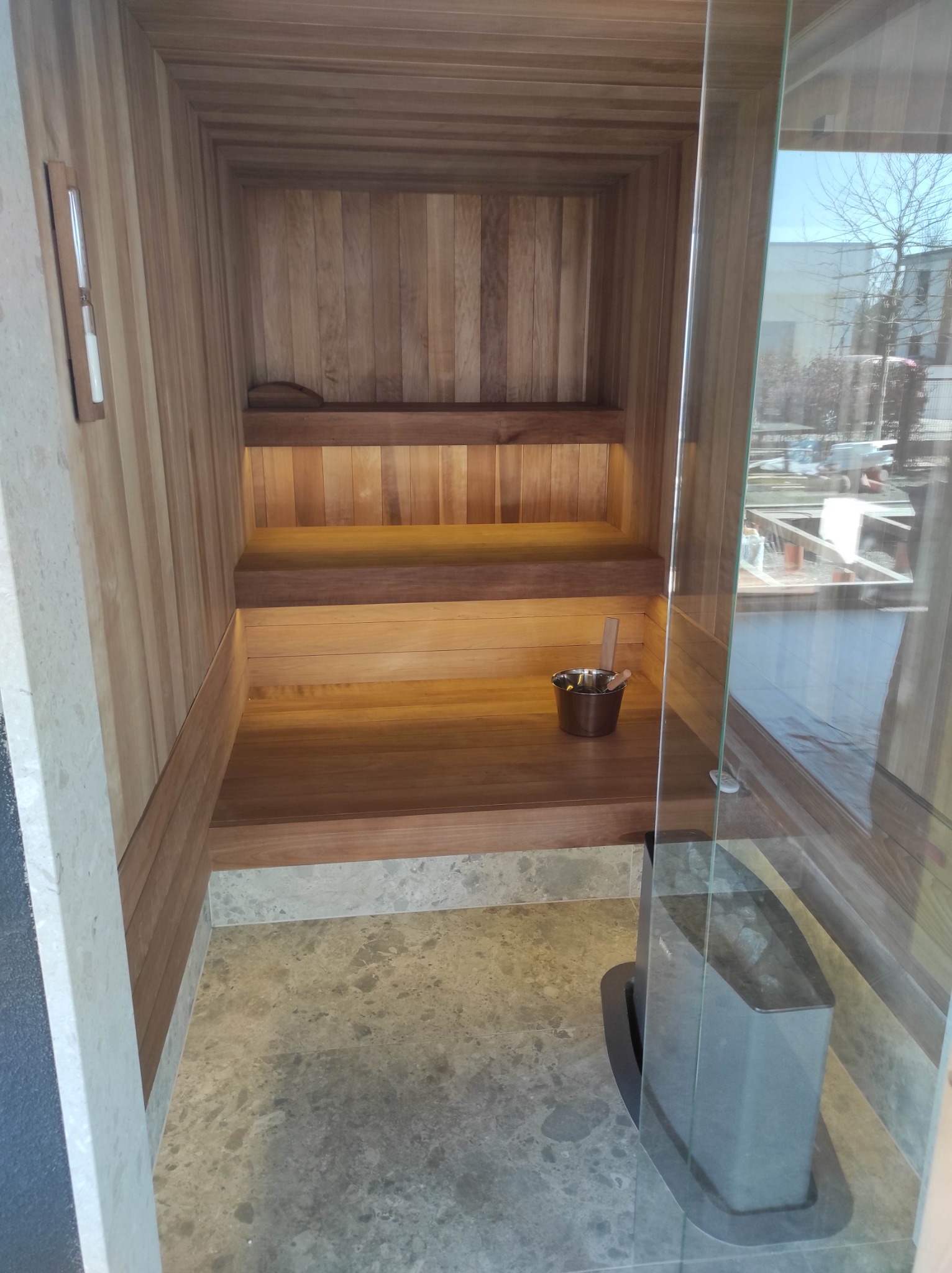 Wnętrze drewnianej kabiny z przeszklonymi drzwiami, widoczny piec z kamieniami, drewniane ławy, klepsydra na ścianie i wiaderko z chochelką.