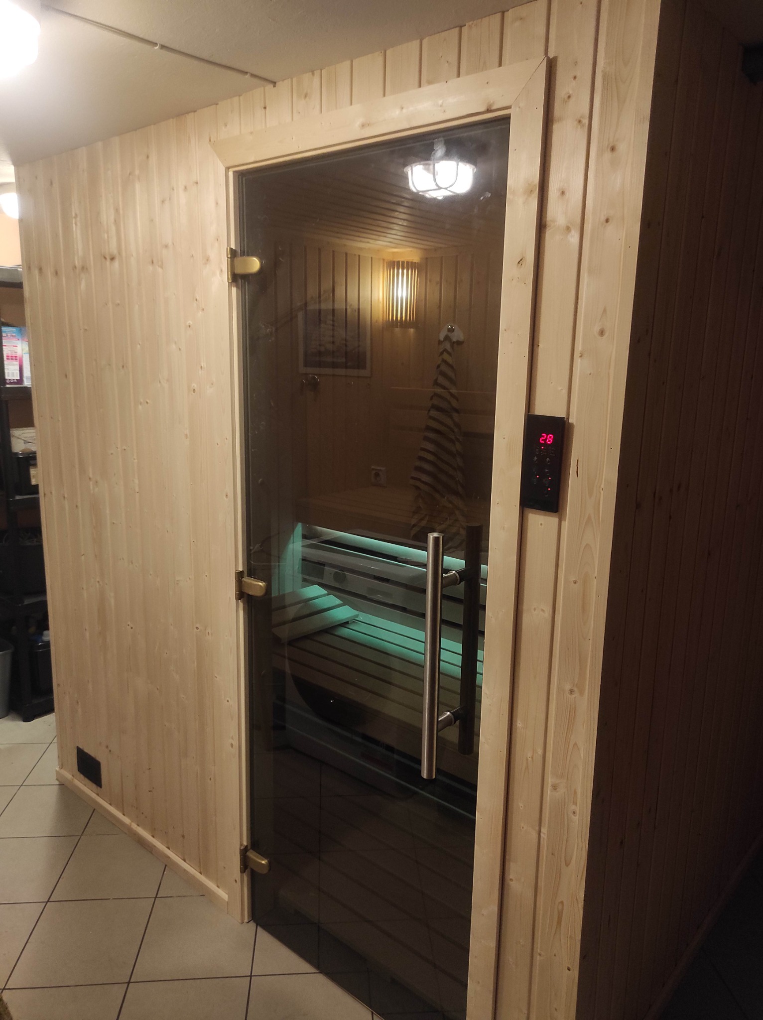 Zamknięta sauna z drewnianymi panelami i szklanymi drzwiami, widoczny panel sterowania z temperaturą 28 stopni Celsjusza i oświetlone wnętrze z leżankami.