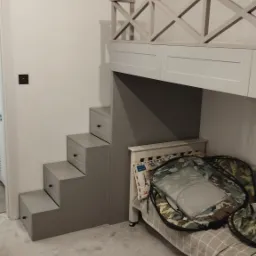 Pokój dziecięcy z łóżkiem na antresoli, do którego prowadzą szare schody z szufladami, zabezpieczone białą, drewnianą balustradą z krzyżowym wzorem.