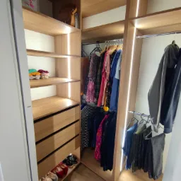 Wnętrze garderoby z oświetleniem LED, drewniane półki, szuflady i wieszaki z ubraniami, widoczne buty na dolnej półce.