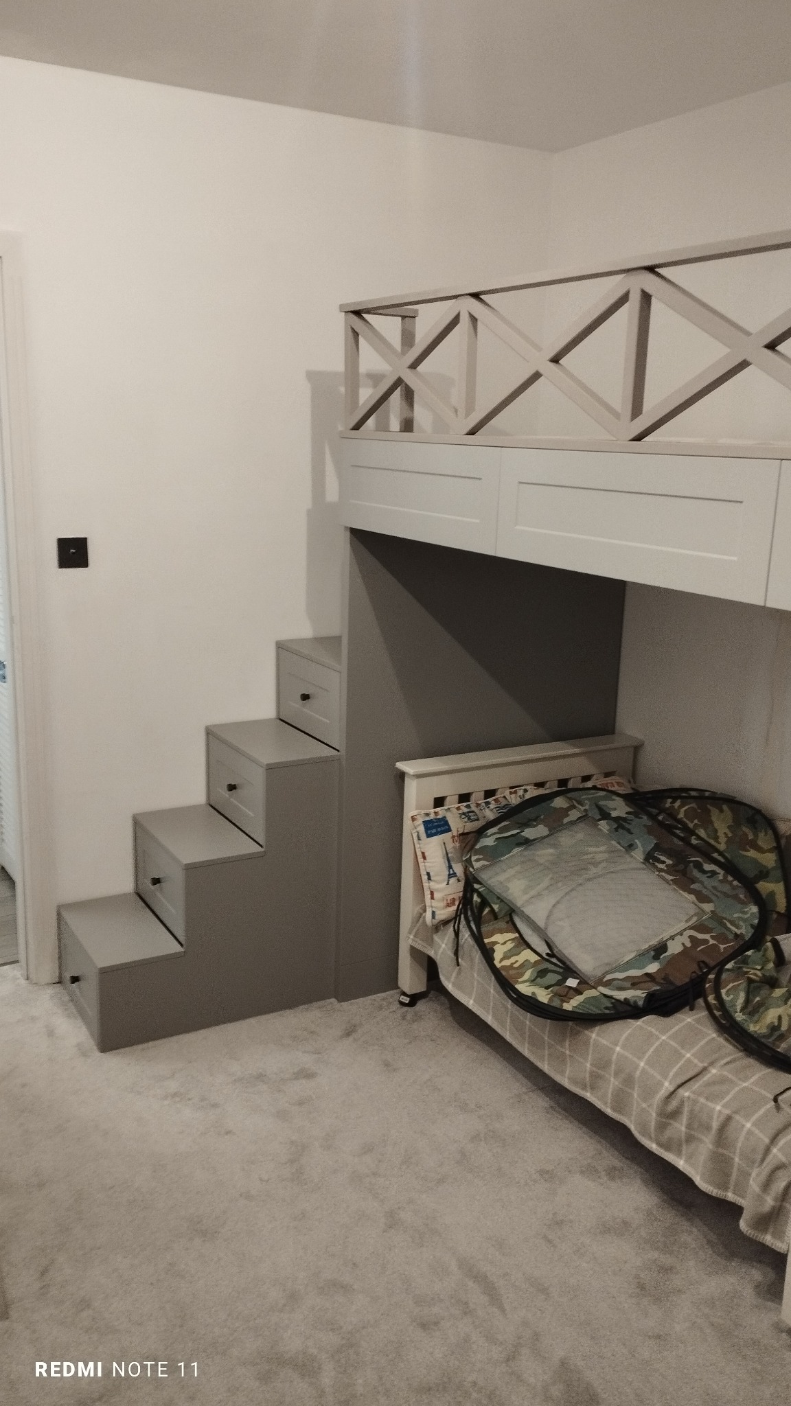 Pokój dziecięcy z łóżkiem na antresoli, do którego prowadzą szare schody z szufladami, zabezpieczone białą, drewnianą balustradą z krzyżowym wzorem.