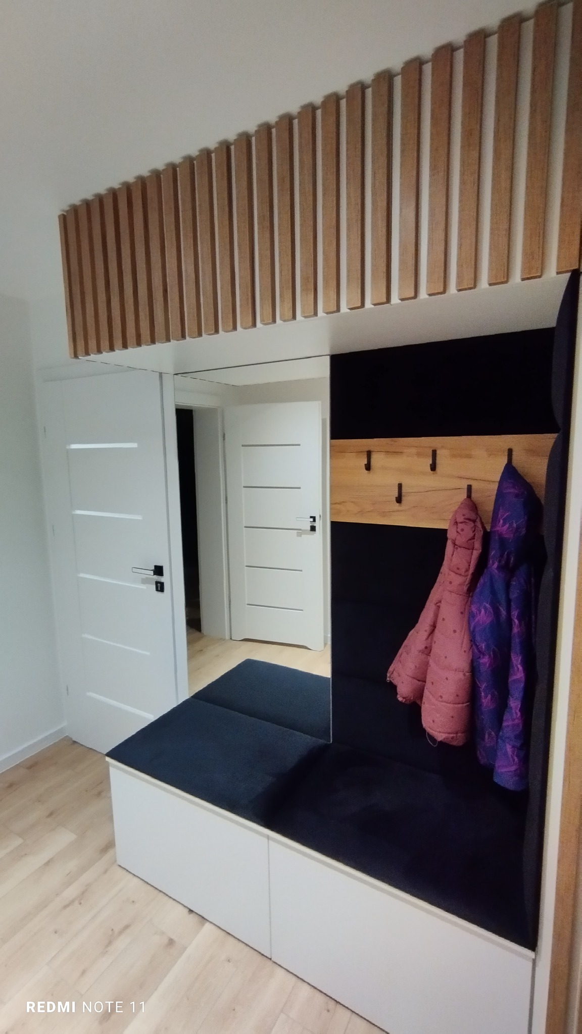 Wnęka w przedpokoju z siedziskiem, wieszakami na ubrania i dekoracyjnymi drewnianymi listwami na suficie, widoczne dwie białe drzwi.