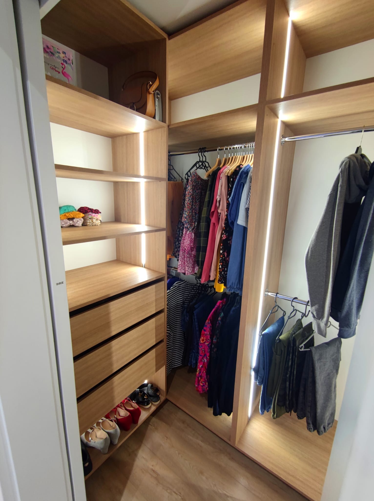 Wnętrze garderoby z oświetleniem LED, drewniane półki, szuflady i wieszaki z ubraniami, widoczne buty na dolnej półce.