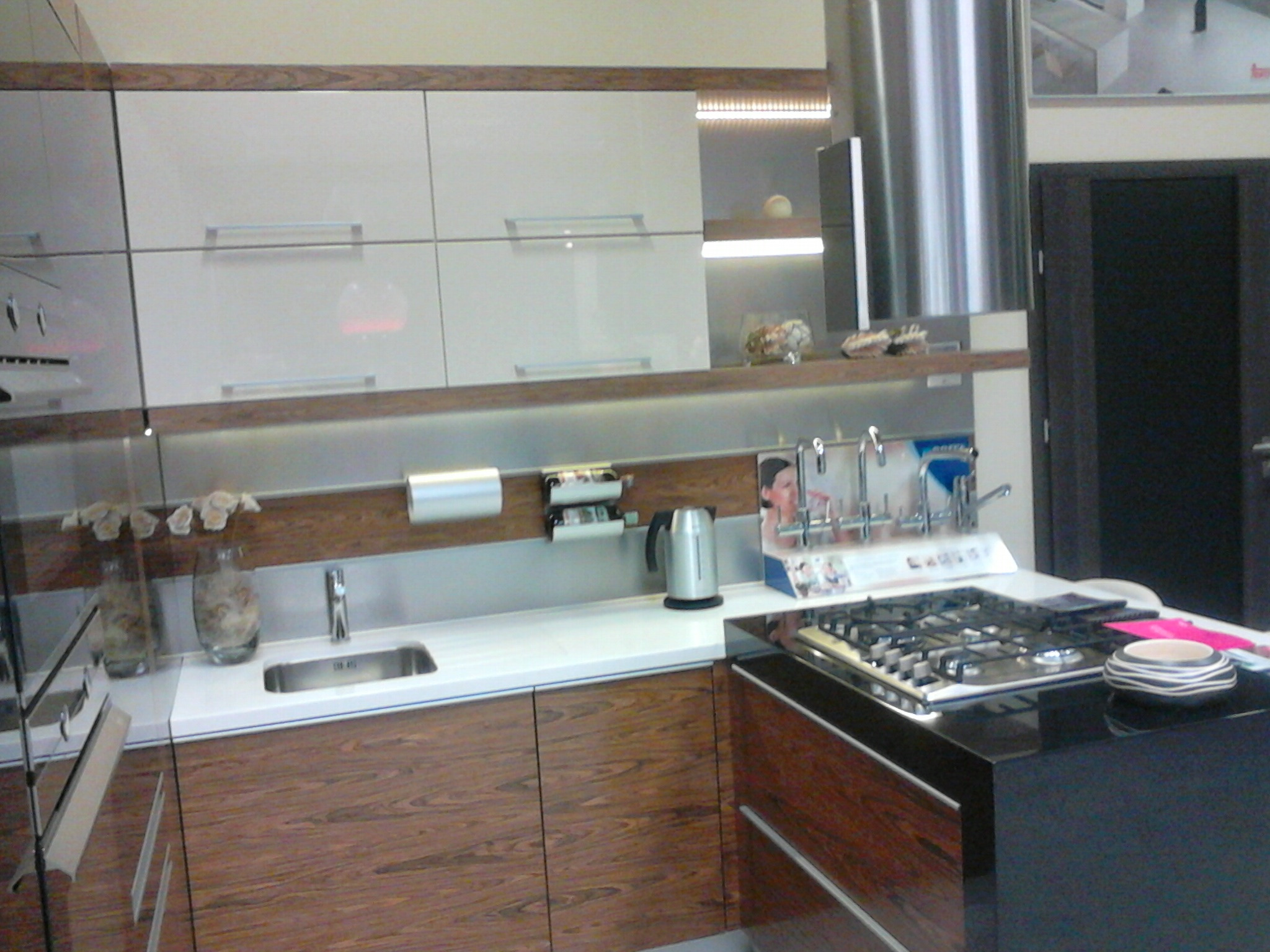 Nowoczesna kuchnia z białymi górnymi szafkami i drewnopodobnymi dolnymi, srebrny okap, czarny blat z kuchenką gazową i dekoracyjną miseczką.