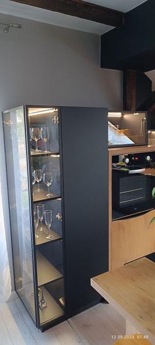 Nowoczesna, czarna szafa witrynowa z przeszklonymi drzwiami i wewnętrznym oświetleniem, eksponująca kieliszki i małą figurkę wieży Eiffla, stanowiąca element zabudowy kuchennej z piekarnikiem...