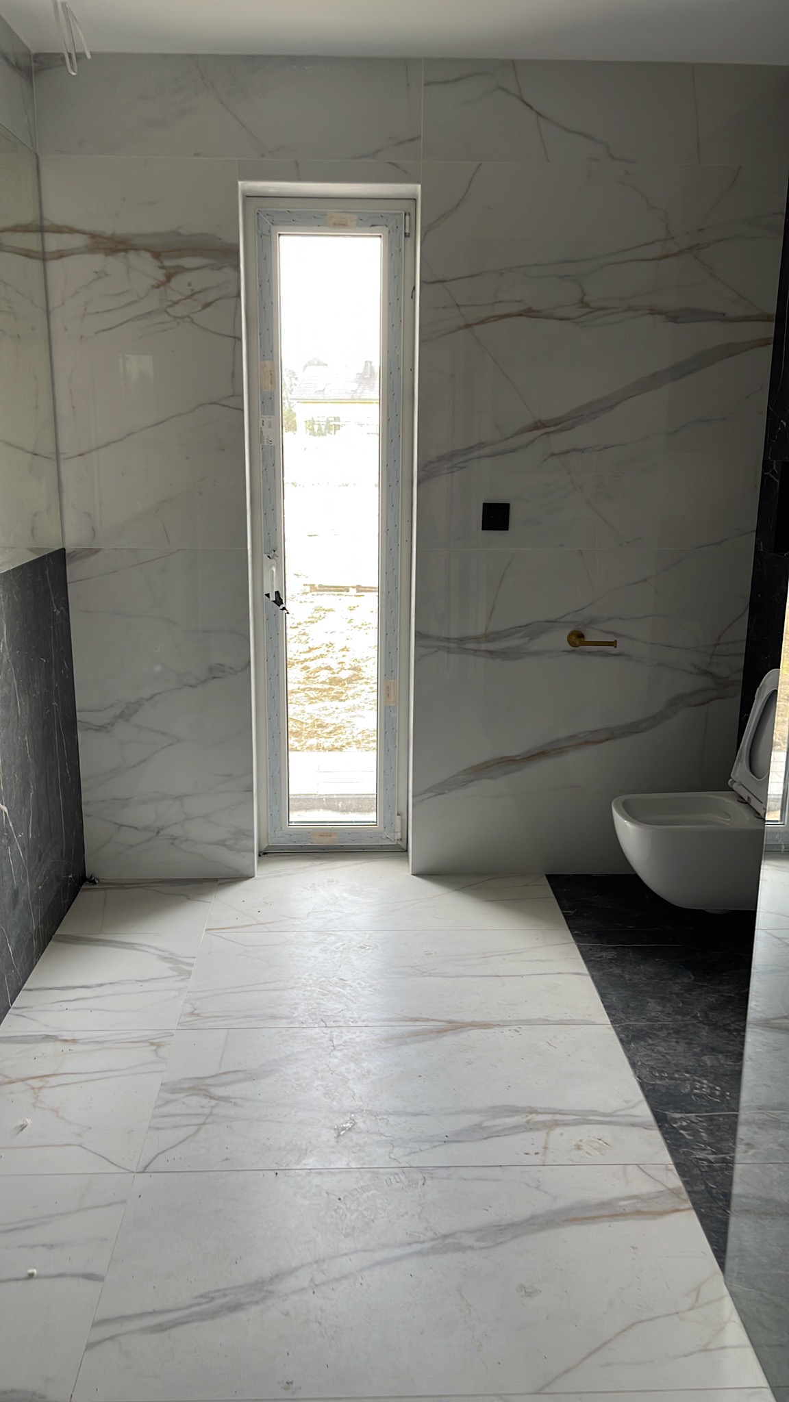 Wykończona łazienka z białymi płytkami imitującymi marmur na ścianach i podłodze, z czarnymi akcentami przy podłodze i w rogu, z toaletą i oknem w tle.